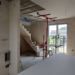 TomDach - Wnętrze w trakcie prac wykończeniowych: ściany przygotowane pod malowanie, częściowo zamontowana płyta na suficie, widoczny podnośnik do płyt i schody.