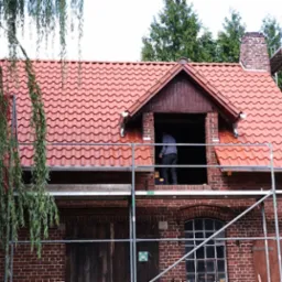 Ceglany budynek z nową, czerwoną dachówką, otoczony rusztowaniami i zielenią. Widoczny fragment pracującego dekarza w otworze dachu.