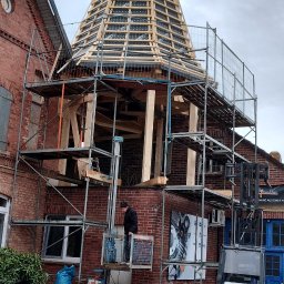 TomDach - Montaż drewnianej konstrukcji dachu wieży na ceglanym budynku, widoczne rusztowania, podnośnik i pracownik w trakcie pracy. Na szczycie dachu wieniec.