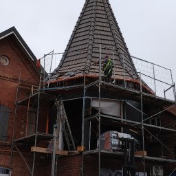 TomDach - Remont wieżyczki z brązową dachówką na ceglanym budynku, otoczonej rusztowaniem. Pracownik w kamizelce odblaskowej i pomarańczowy wózek widłowy.