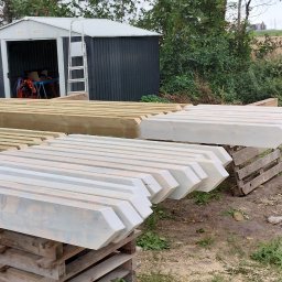 TomDach - Przygotowane elementy więźby dachowej, część pomalowana na biało, leżące na paletach i drewnianych kozłach, w tle budynek w trakcie budowy i zielona roślinność.