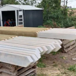 Przygotowane elementy więźby dachowej, część pomalowana na biało, leżące na paletach i drewnianych kozłach, w tle budynek w trakcie budowy i zielona roślinność.