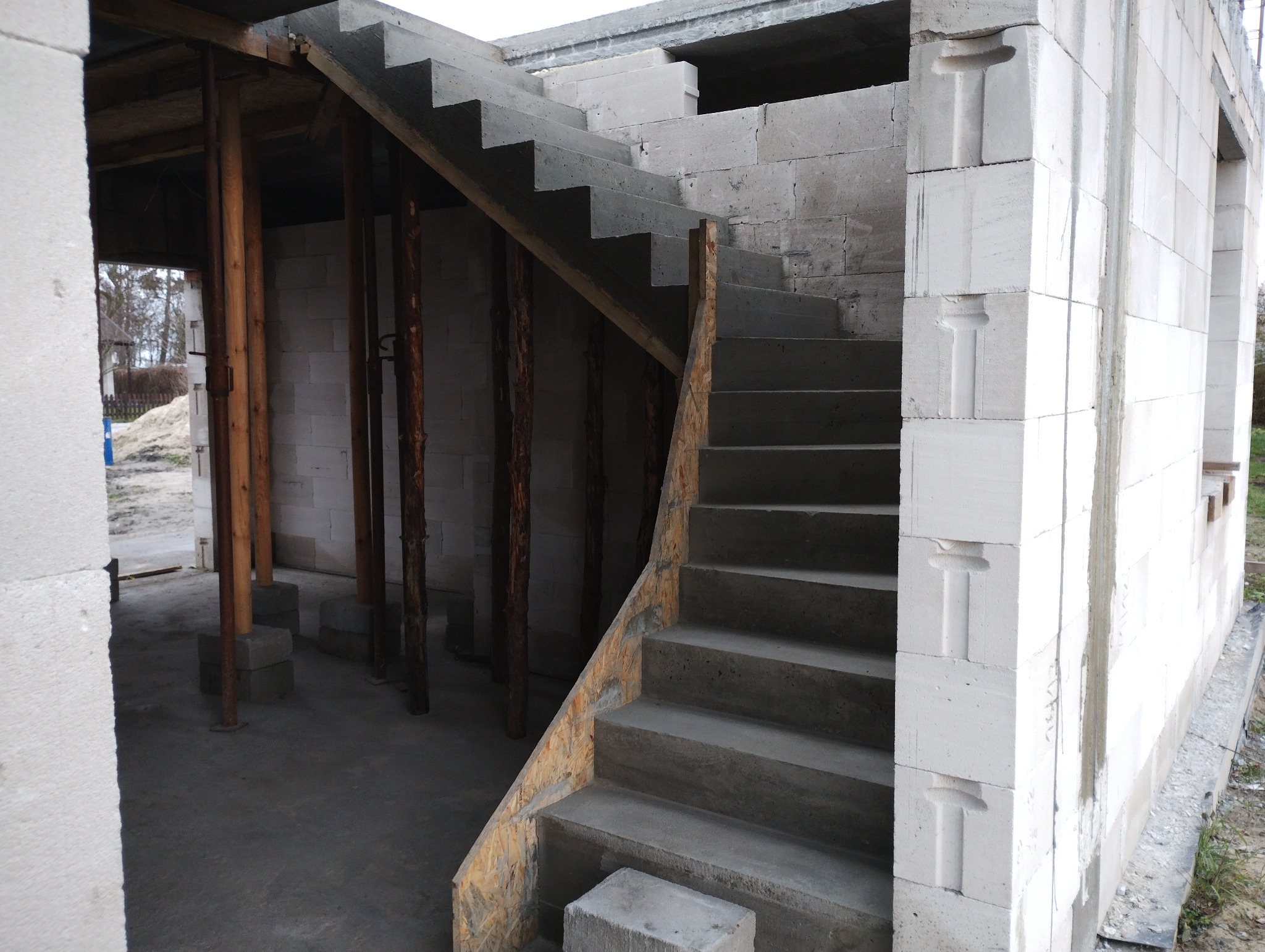 Wnętrze budynku w budowie: betonowe schody z drewnianym szalunkiem, ściany z betonu komórkowego z charakterystycznymi wcięciami, drewniane podpory stropu.