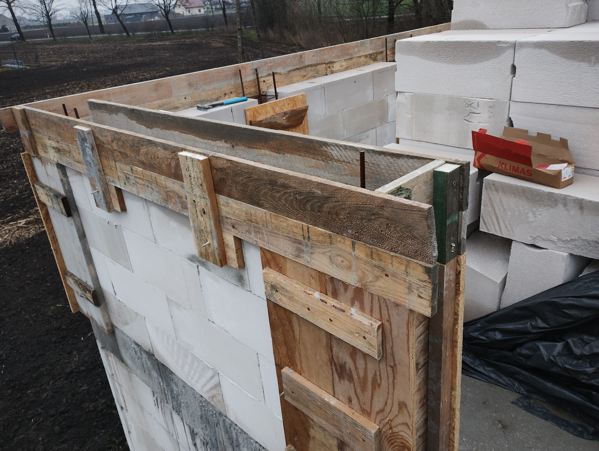 Budowa ściany z bloczków betonu komórkowego Ytong, z drewnianym szalunkiem i widocznymi narzędziami. W tle ziemia i zabudowania.