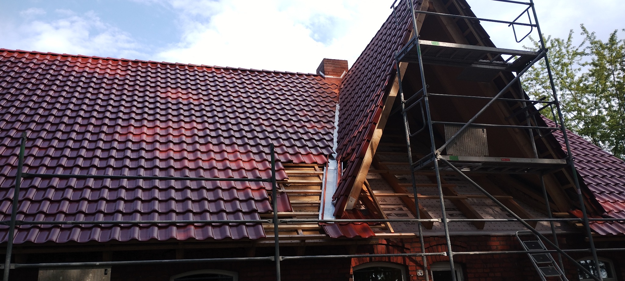 Częściowo zdjęte czerwone dachówki na budynku z cegły, widoczne rusztowanie i elementy więźby. Etap wymiany pokrycia dachowego w słoneczny dzień.