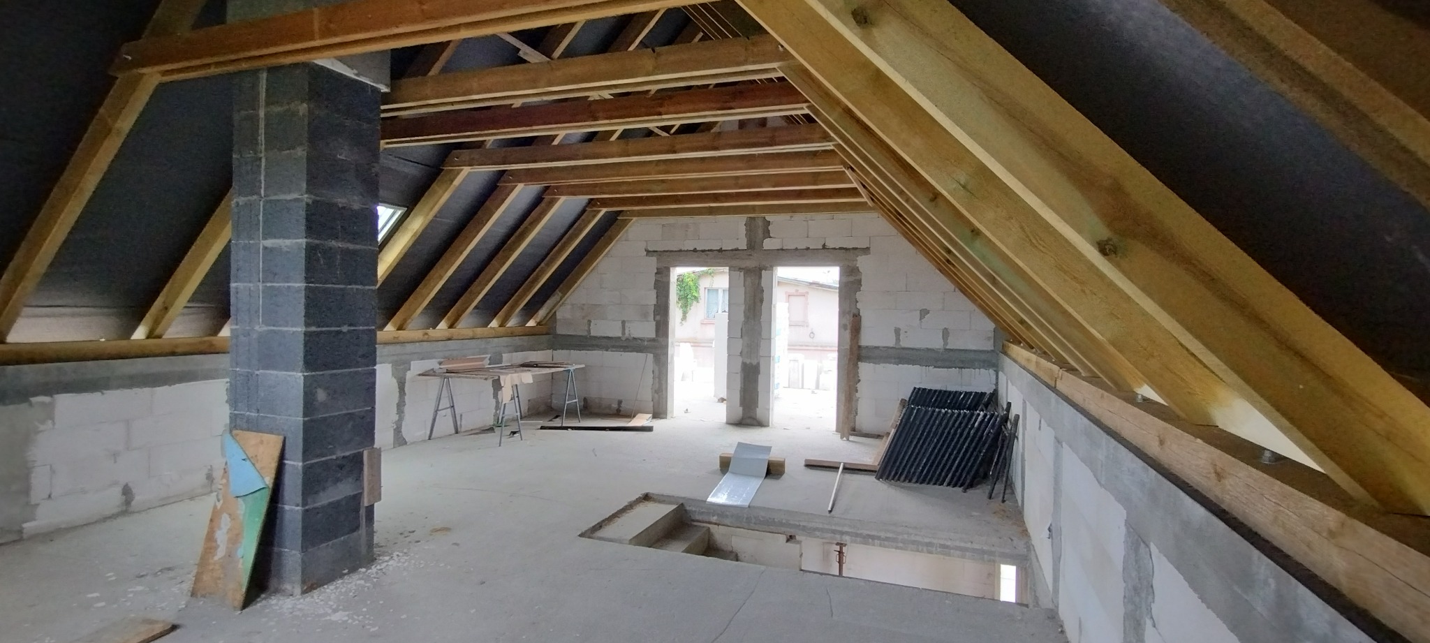 Poddasze w trakcie adaptacji: widoczna więźba dachowa, ściany z bloczków, kolumna z cegły. Częściowo wykończone wnętrze z oknami dachowymi i wyjściem na zewnątrz.