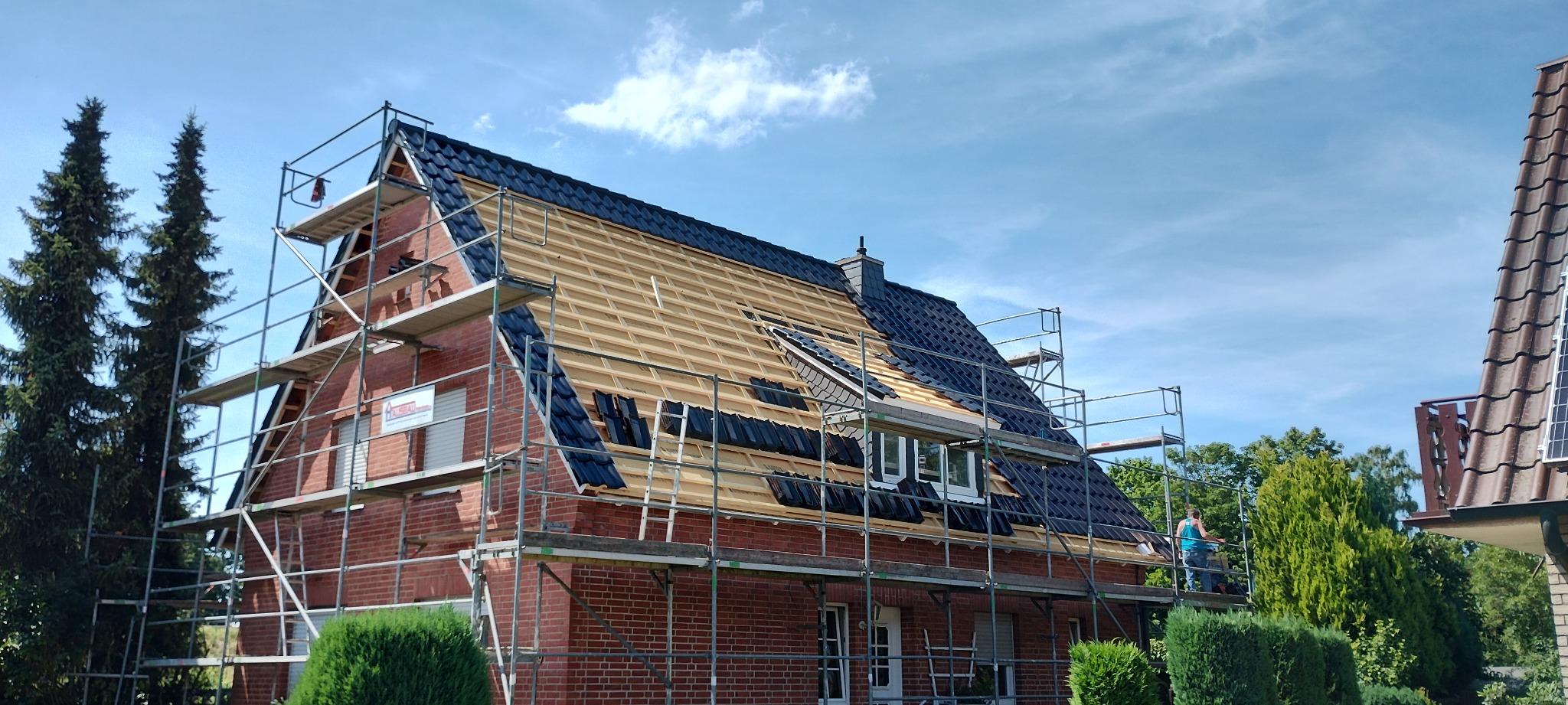 Ceglany dom w rusztowaniach, częściowo pokryty czarną dachówką. Widoczna drewniana konstrukcja dachu i pracownik na rusztowaniu. Błękitne niebo w tle.