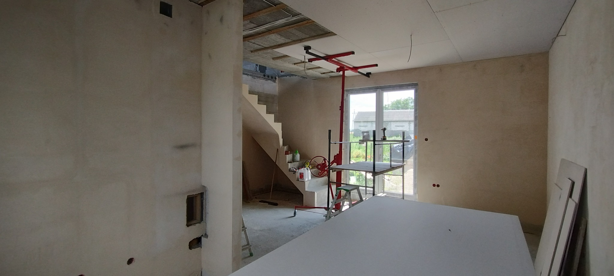 Wnętrze w trakcie prac wykończeniowych: ściany przygotowane pod malowanie, częściowo zamontowana płyta na suficie, widoczny podnośnik do płyt i schody.