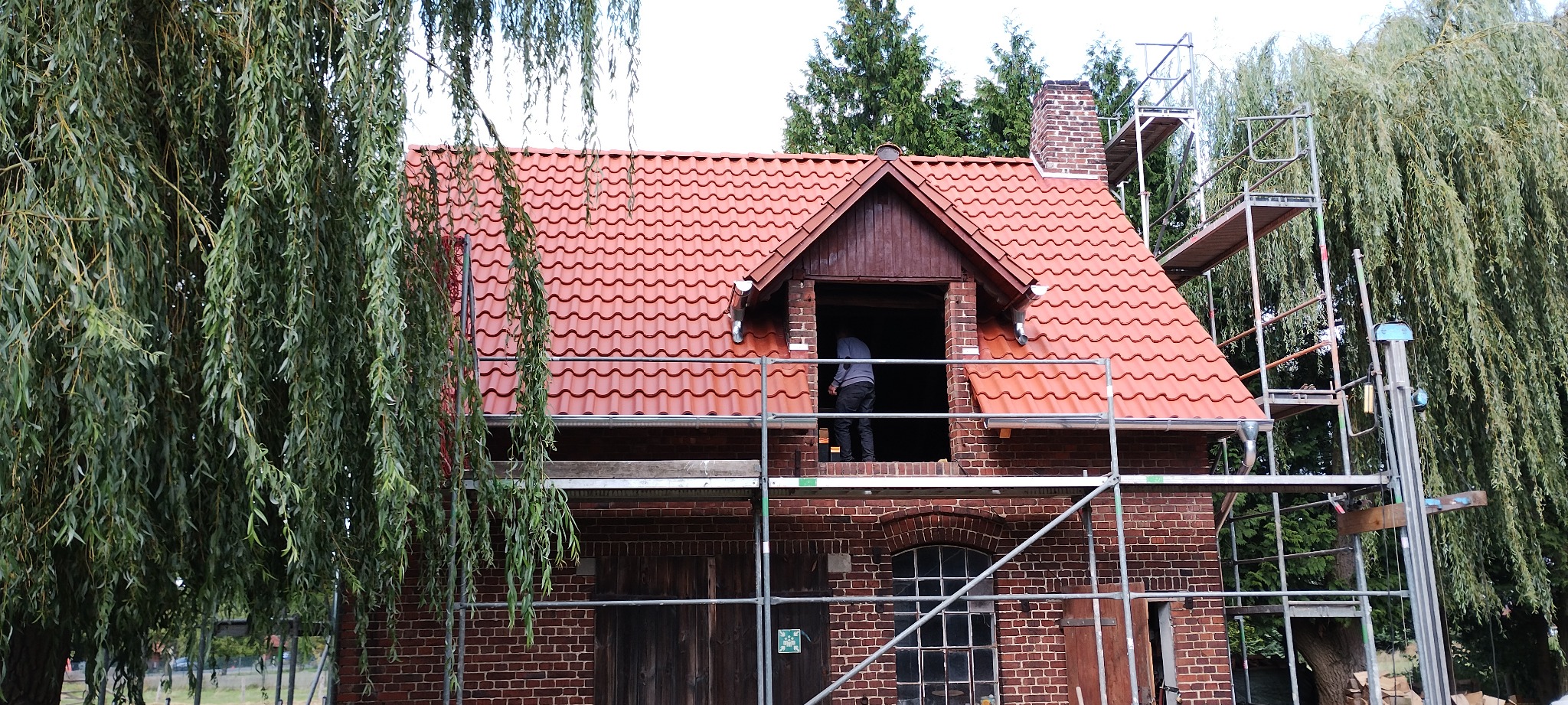 Ceglany budynek z nową, czerwoną dachówką, otoczony rusztowaniami i zielenią. Widoczny fragment pracującego dekarza w otworze dachu.