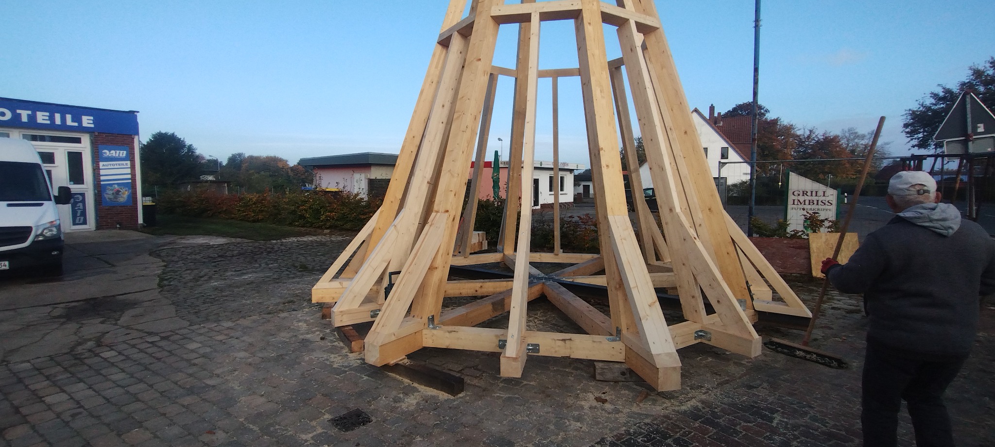 Wznoszenie drewnianej konstrukcji o stożkowym kształcie, prawdopodobnie elementu większej budowli. Widoczne złączenia i surowe drewno. W tle budynki i zieleń.