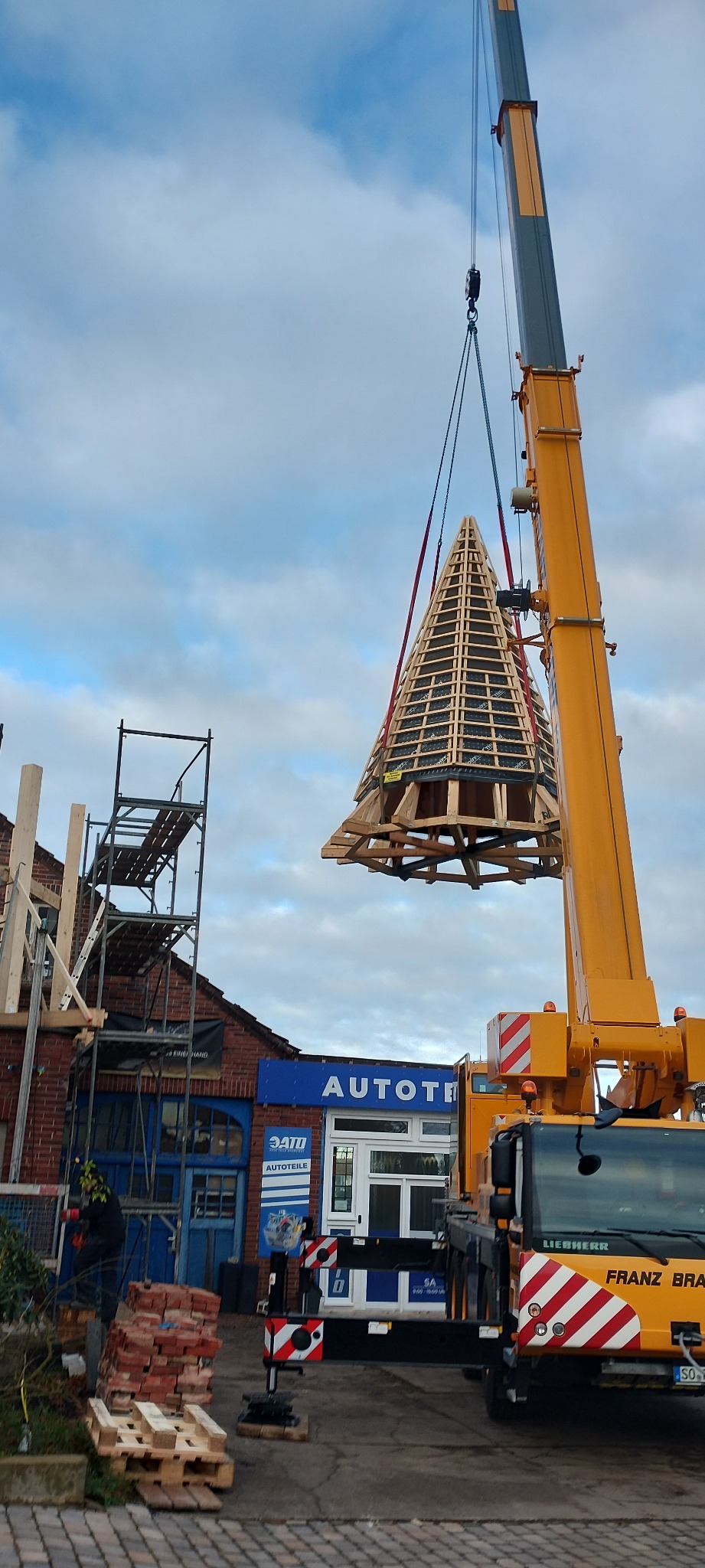 Montaż wieżyczki dachowej z drewnianą konstrukcją za pomocą dźwigu Liebherr, na tle budynku z cegły i błękitnego nieba.