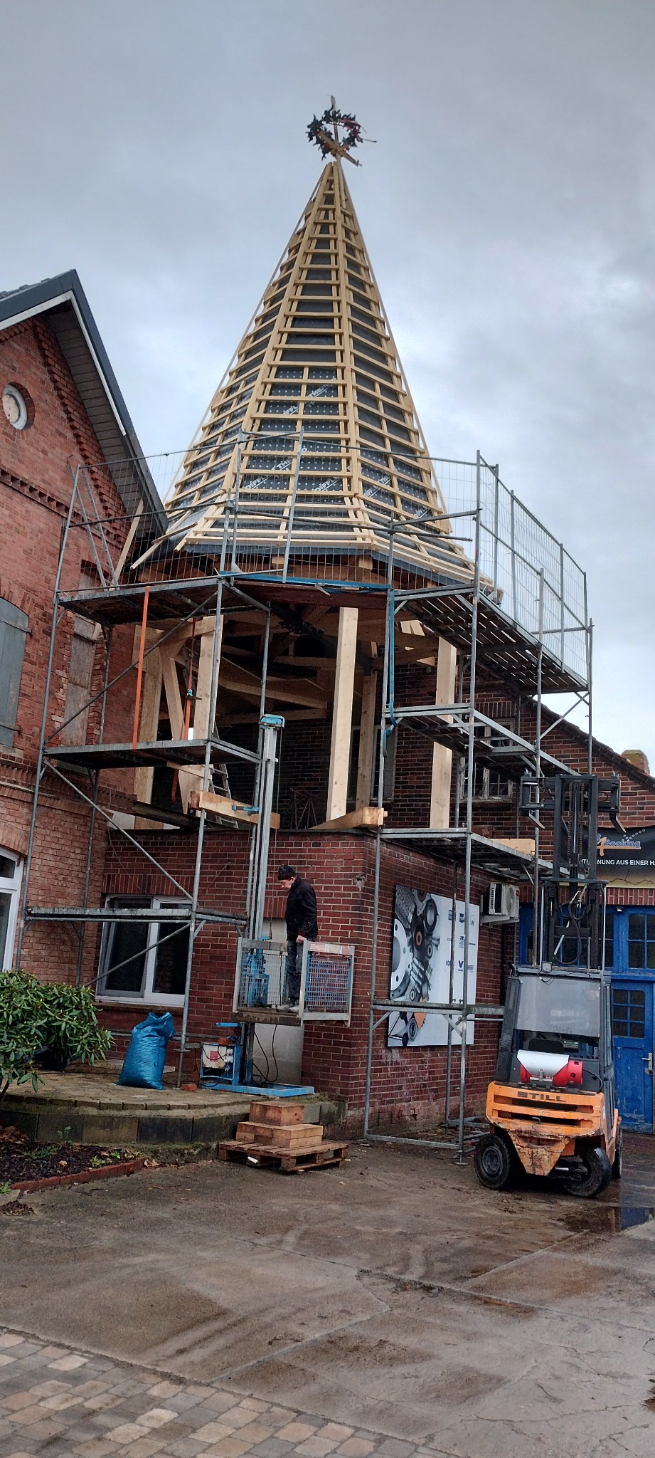 Montaż drewnianej konstrukcji dachu wieży na ceglanym budynku, widoczne rusztowania, podnośnik i pracownik w trakcie pracy. Na szczycie dachu wieniec.