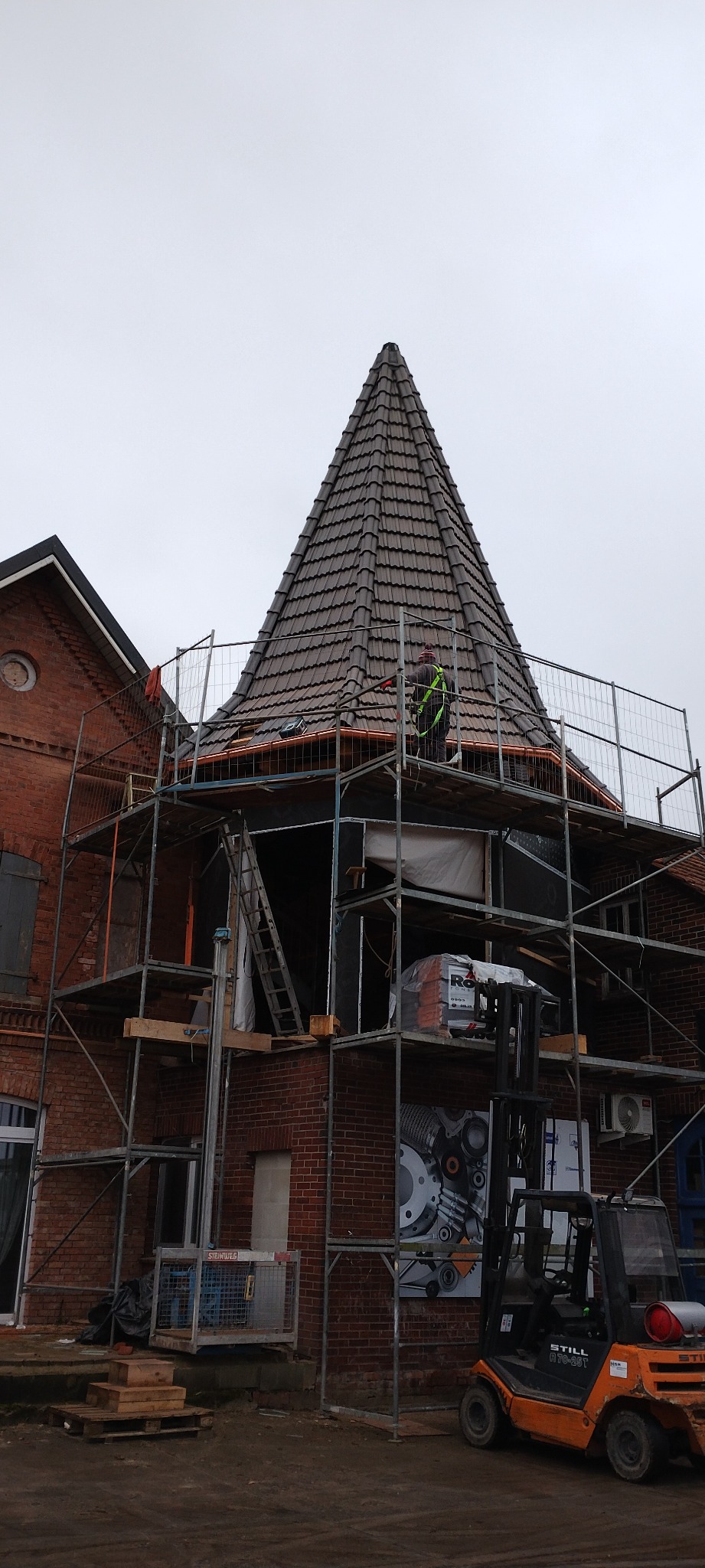 Remont wieżyczki z brązową dachówką na ceglanym budynku, otoczonej rusztowaniem. Pracownik w kamizelce odblaskowej i pomarańczowy wózek widłowy.