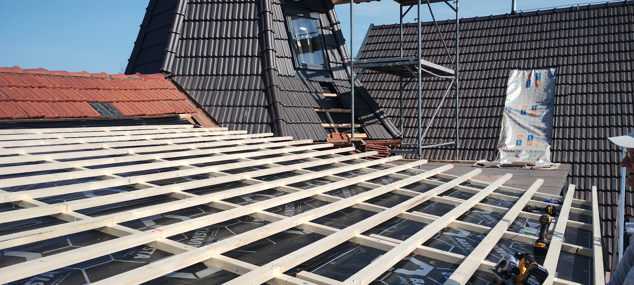 Wysoki kąt: montaż drewnianej konstrukcji na dachu pokrytym czarną membraną, w tle fragmenty dachów z dachówką w odcieniach czerwieni i grafitu oraz rusztowanie.