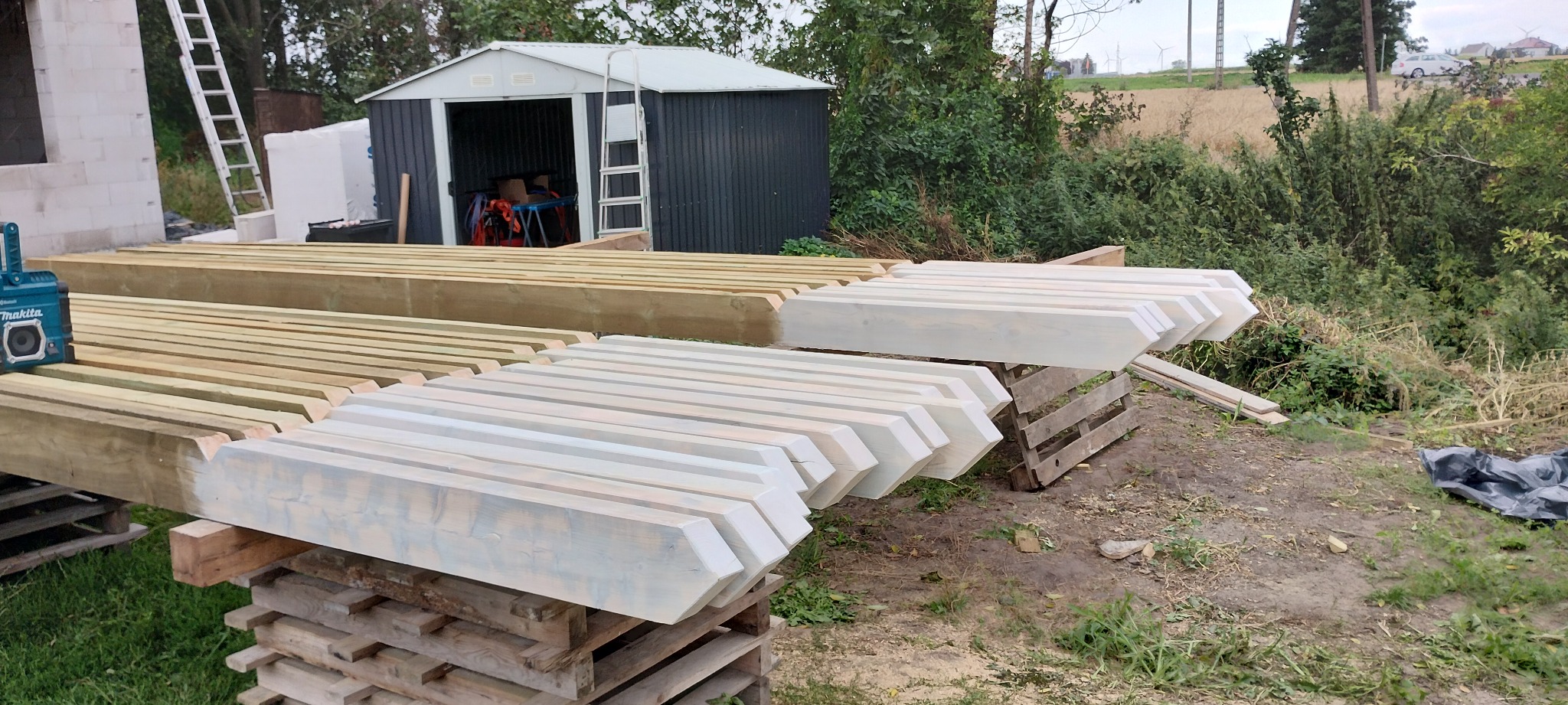 Przygotowane elementy więźby dachowej, część pomalowana na biało, leżące na paletach i drewnianych kozłach, w tle budynek w trakcie budowy i zielona roślinność.