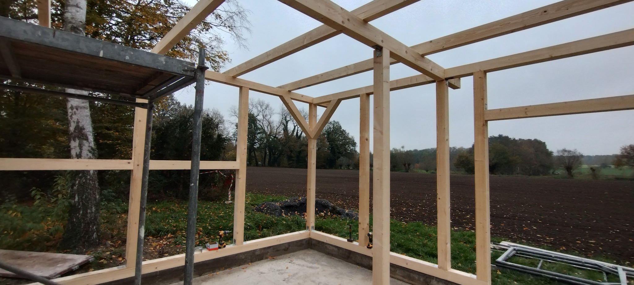 Wznoszenie drewnianej konstrukcji szkieletowej w Inowrocławiu, widok na pole. Elementy więźby dachowej połączone metalowymi złączkami. Na pierwszym planie rusztowanie.