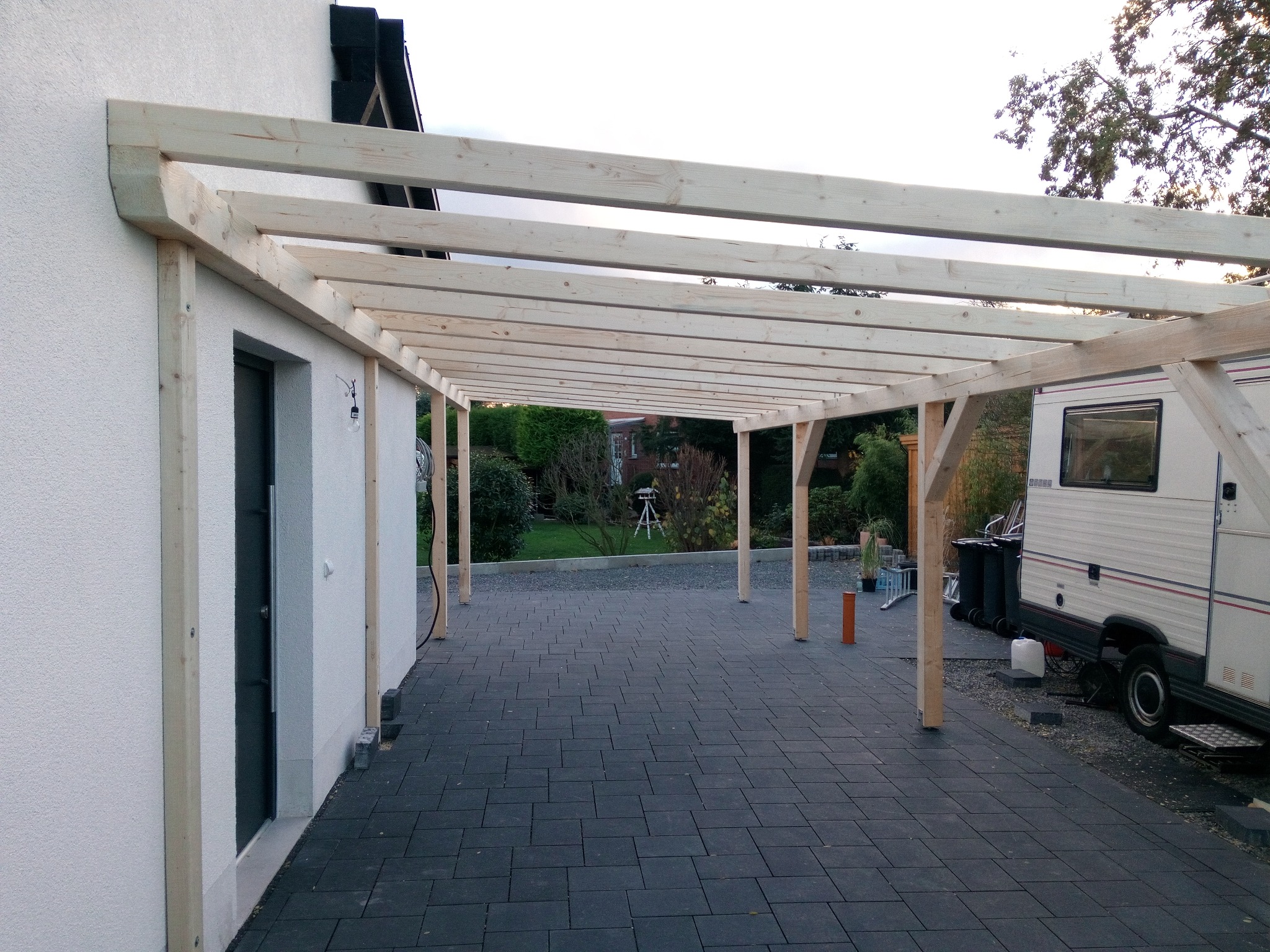 Nowa, surowa więźba drewniana z jasnego drewna tworząca zadaszenie tarasu przy białym budynku. Widok z boku na konstrukcję i szarą kostkę brukową.