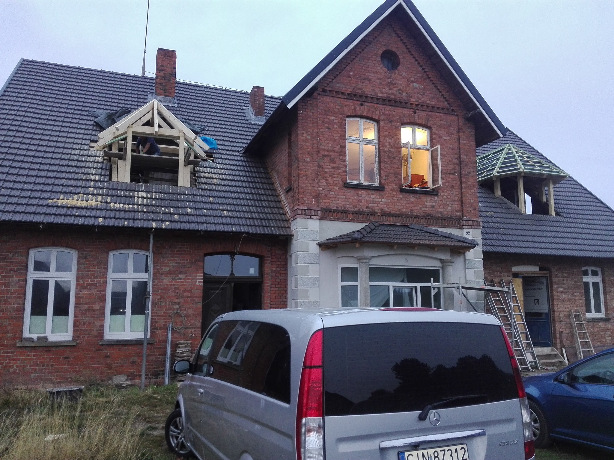 Ceglany budynek w trakcie prac dekarskich: montaż więźby dachowej lukarny i konstrukcji dachu nad wejściem. Na pierwszym planie srebrny samochód.