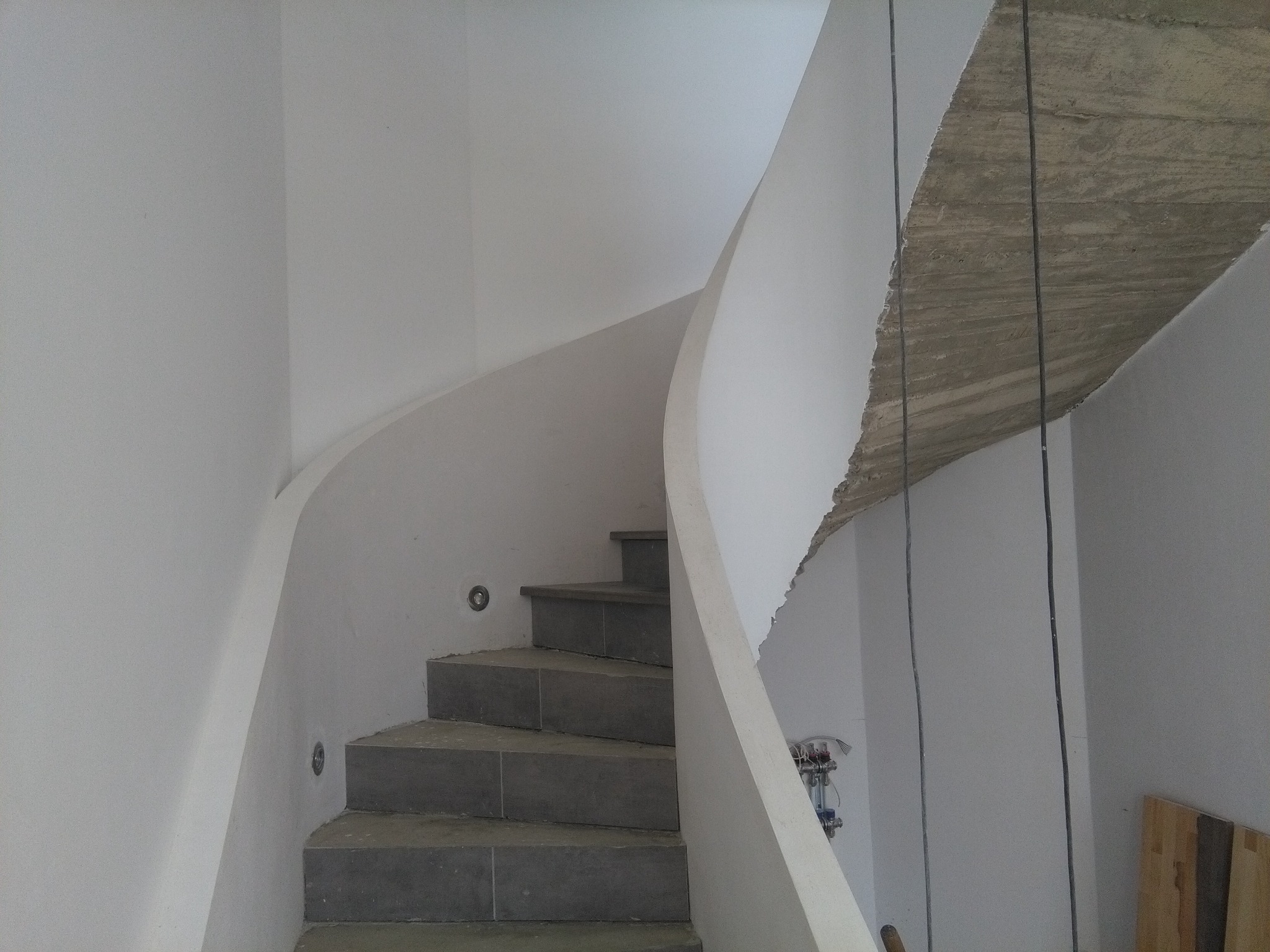 Wykończone, spiralne schody z szarymi stopniami i oświetleniem punktowym w białej klatce schodowej. Surowy betonowy strop w tle. Etap prac wykończeniowych.