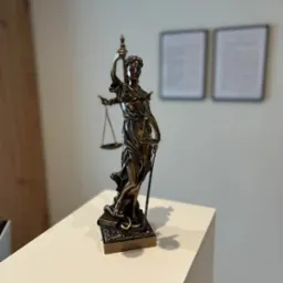 Statua Temidy z wagą i mieczem, symbol sprawiedliwości, na białym biurku. W tle dwa dyplomy w ramkach.