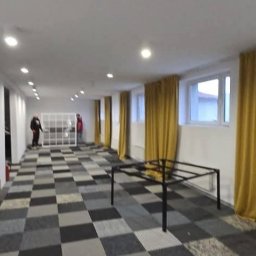 INTERPODŁOGA Marek Wyciszkiewicz - Wnętrze po montażu wykładziny dywanowej w szachownicę, z żółtymi zasłonami i osobami w tle. Widoczne metalowe stelaże mebli.