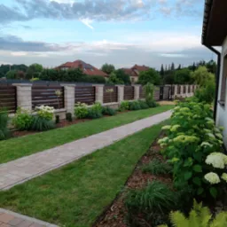 Uporządkowany ogród z ścieżką z kostki, rabatami z korą, hortensjami i ozdobnym ogrodzeniem z drewnianymi panelami w słoneczny dzień.