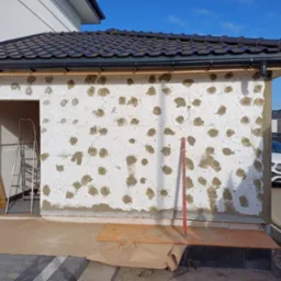 Ocieplanie ściany zewnętrznej budynku z widoczną warstwą izolacyjną i przygotowanym podłożem pod tynk. Widoczny fragment dachu i rynny.