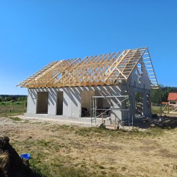 NordHouse MAŁGORZATA KOSMALSKA - Surowy dom z betonu komórkowego w trakcie budowy, z drewnianą konstrukcją dachu w słoneczny dzień. Widoczne rusztowania i teren budowy.