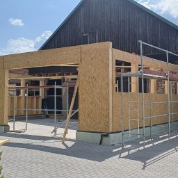 NordHouse MAŁGORZATA KOSMALSKA - Budowa szkieletowa budynku w trakcie realizacji, z widocznym drewnianym szkieletem i płytami OSB, rusztowaniem oraz elementami konstrukcyjnymi na zewnątrz.