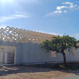 NordHouse MAŁGORZATA KOSMALSKA - Wznoszenie więźby dachowej na budynku z bloczków betonu komórkowego, widok z zewnątrz, drzewo w pobliżu, ciężarówka w tle, słoneczny dzień.