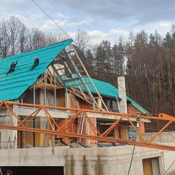 NordHouse MAŁGORZATA KOSMALSKA - Budowa domu jednorodzinnego z zielonym dachem w trakcie realizacji. Na dachu dwóch pracowników. Na pierwszym planie pomarańczowy dźwig budowlany.