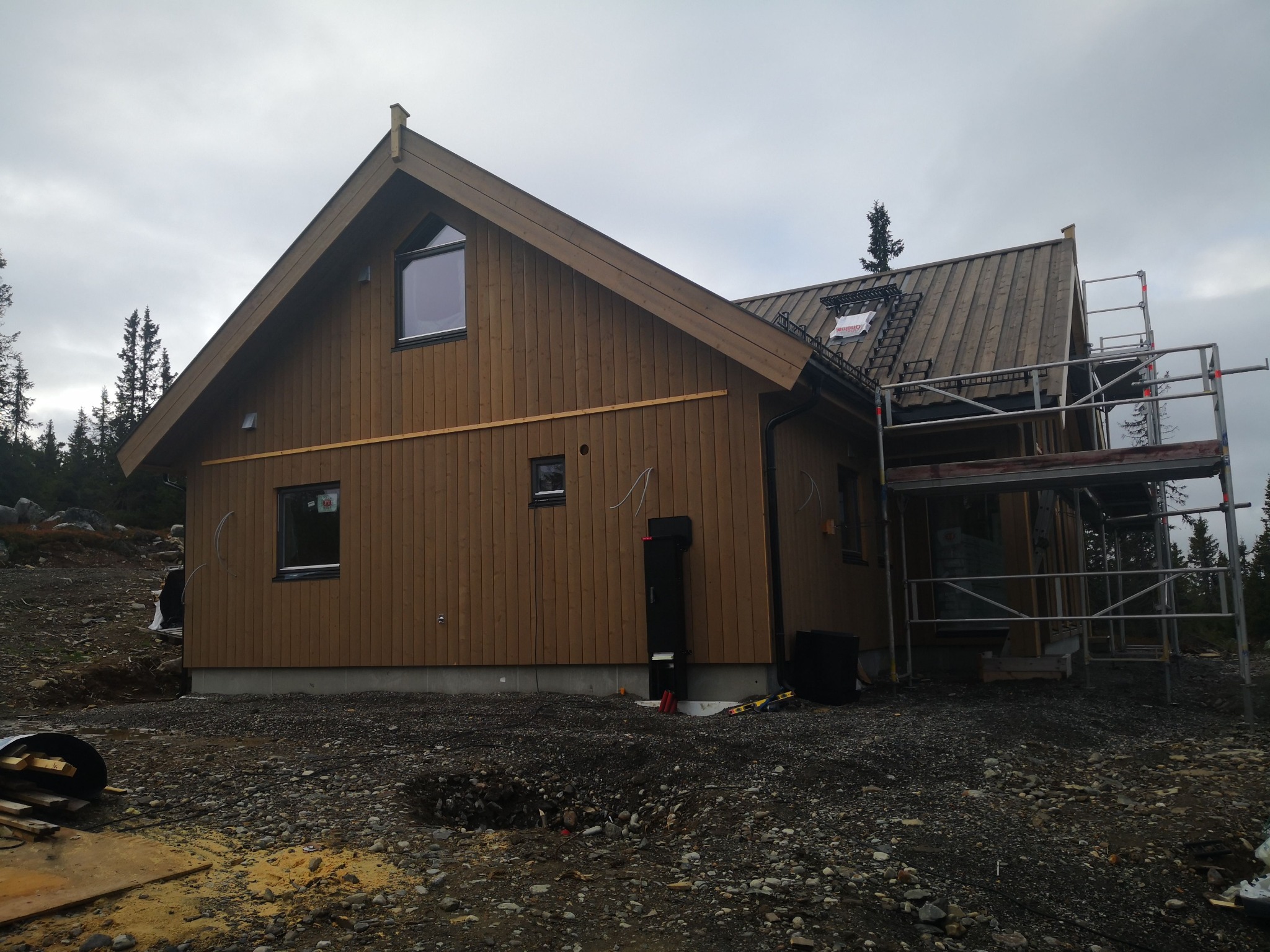 Dom w trakcie budowy z drewnianą elewacją, widoczne rusztowania i elementy dachu. Surowy teren wokół budynku, pochmurne niebo w tle.