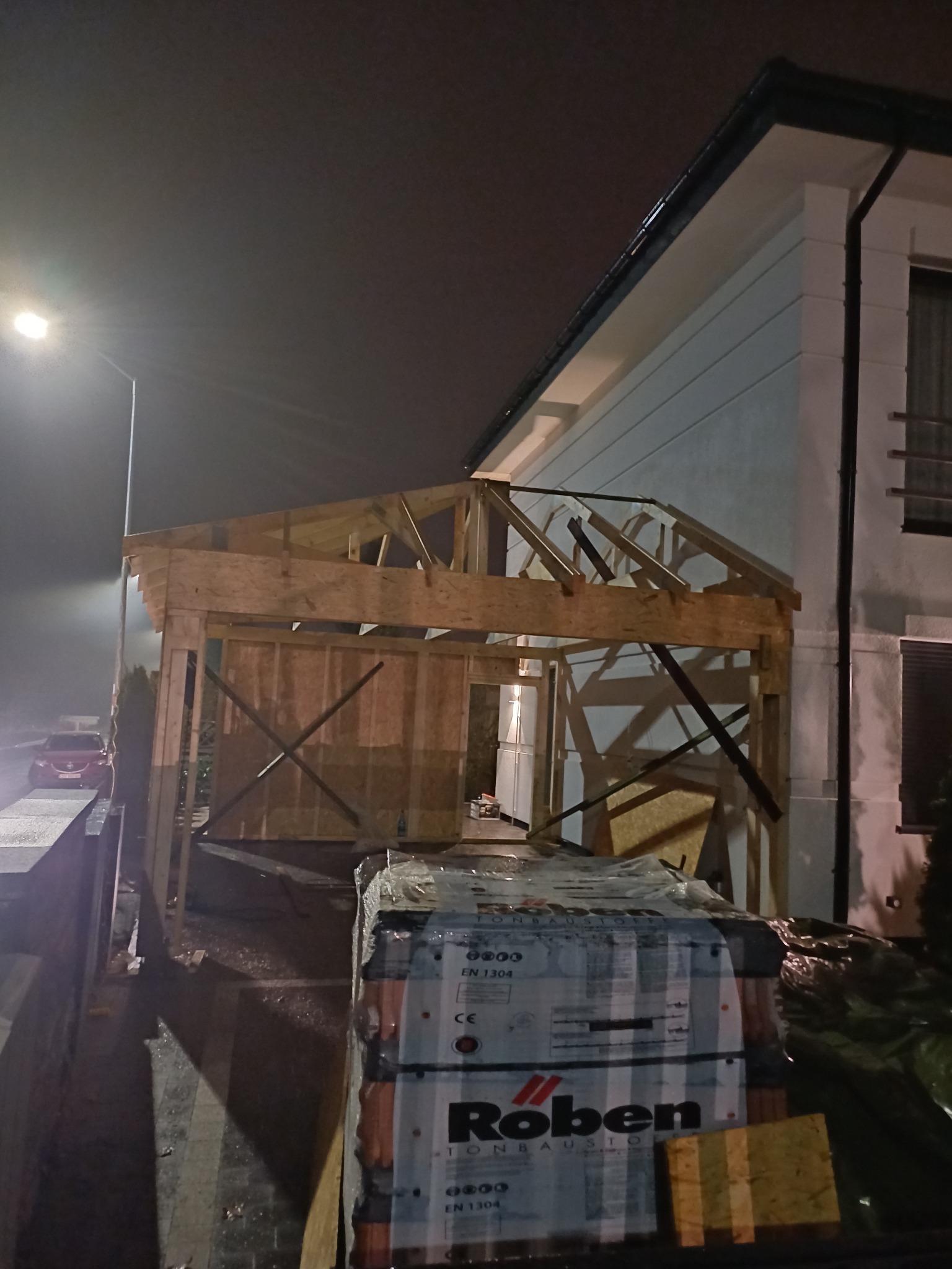 W trakcie budowy drewnianej konstrukcji dachu przy domu jednorodzinnym, widoczne pakiety dachówek Roben, oświetlone nocą lampą uliczną.