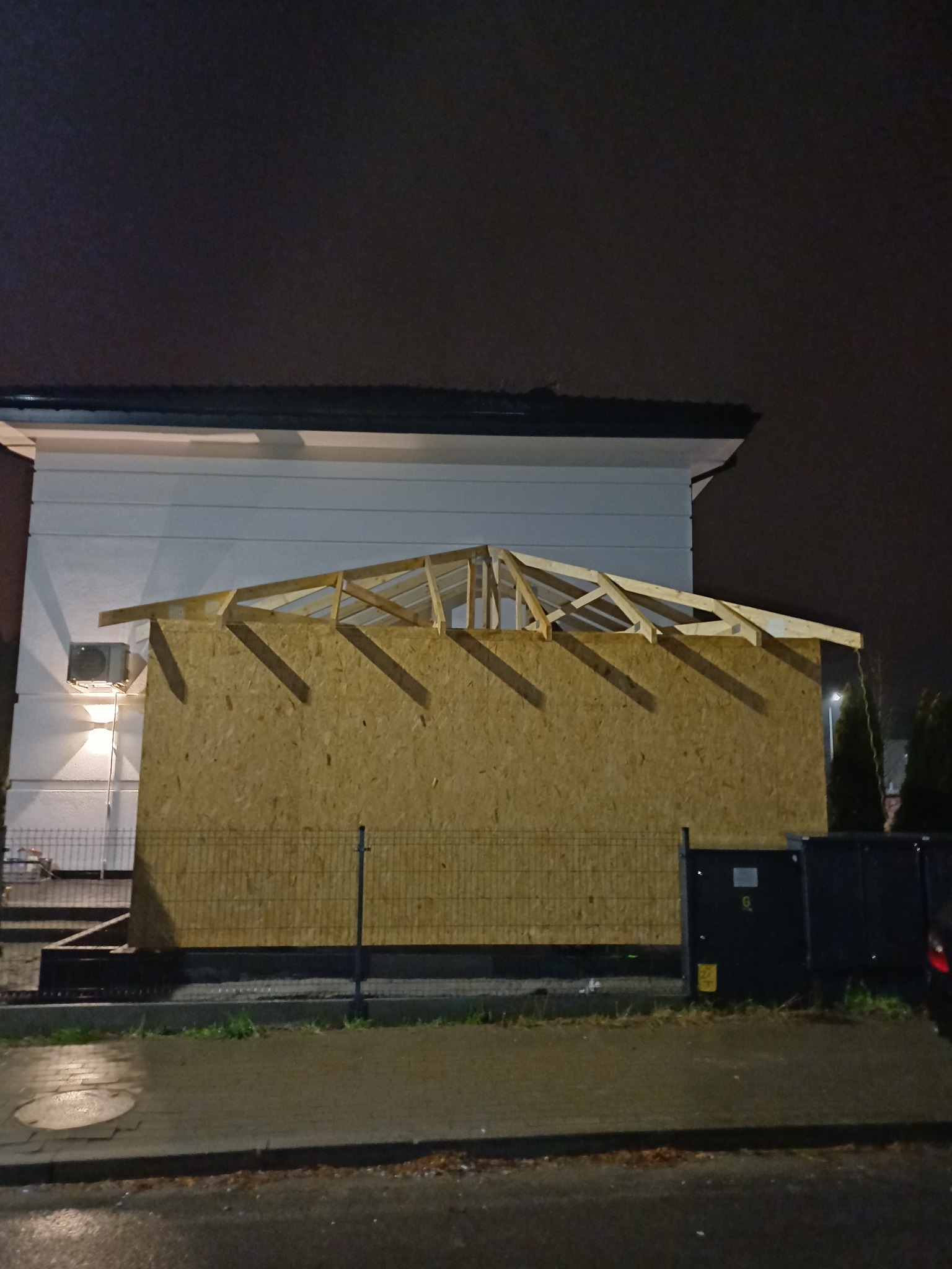 Nocne ujęcie budynku z widoczną konstrukcją dachu w trakcie budowy, z deskami OSB i drewnianą więźbą, oświetlone sztucznym światłem.