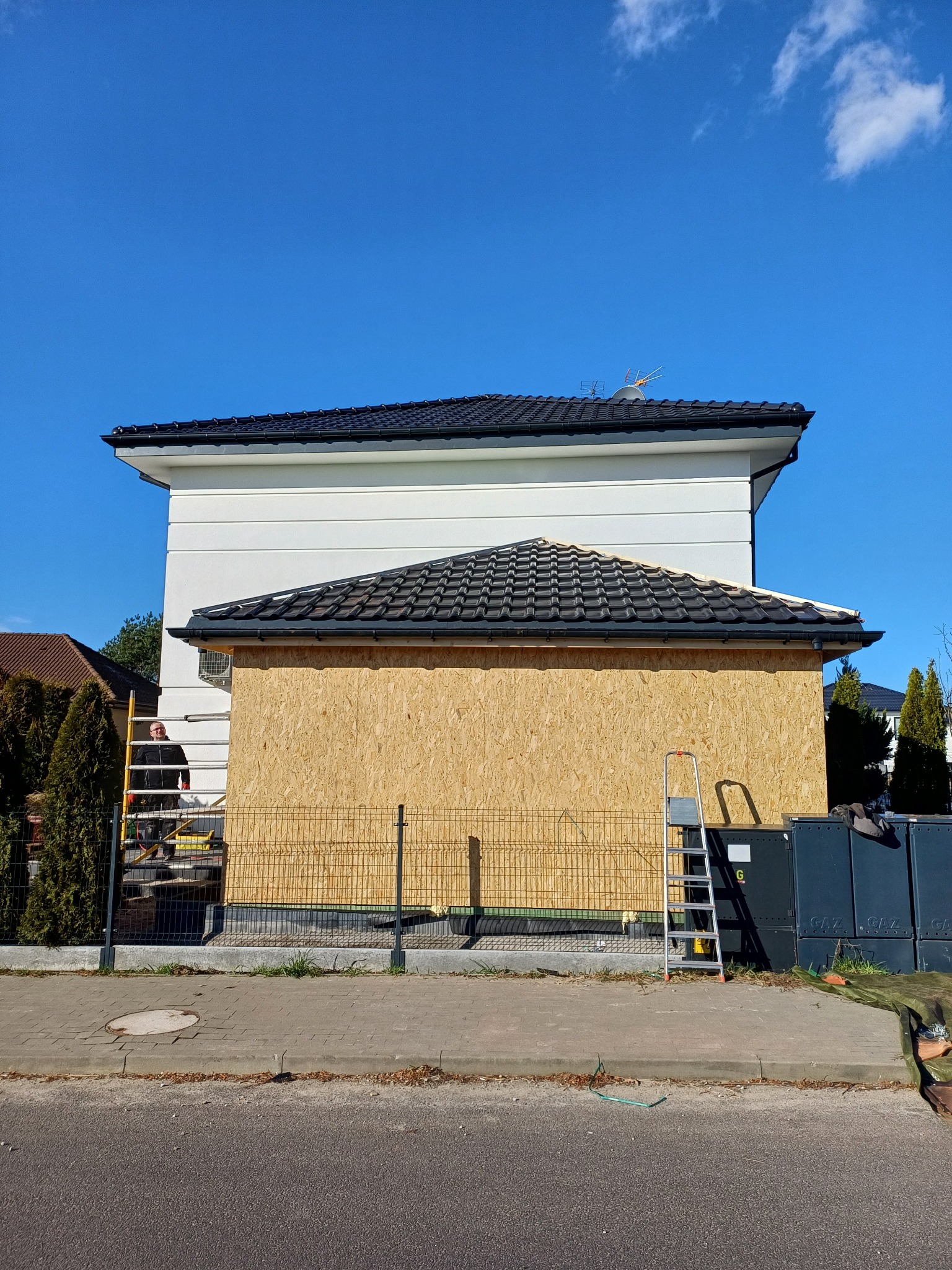 Dom w trakcie prac dekarskich, widoczna drewniana konstrukcja dachu i drabina. Na rusztowaniu stoi dekarz. Dach pokryty czarną dachówką, błękitne niebo w tle.