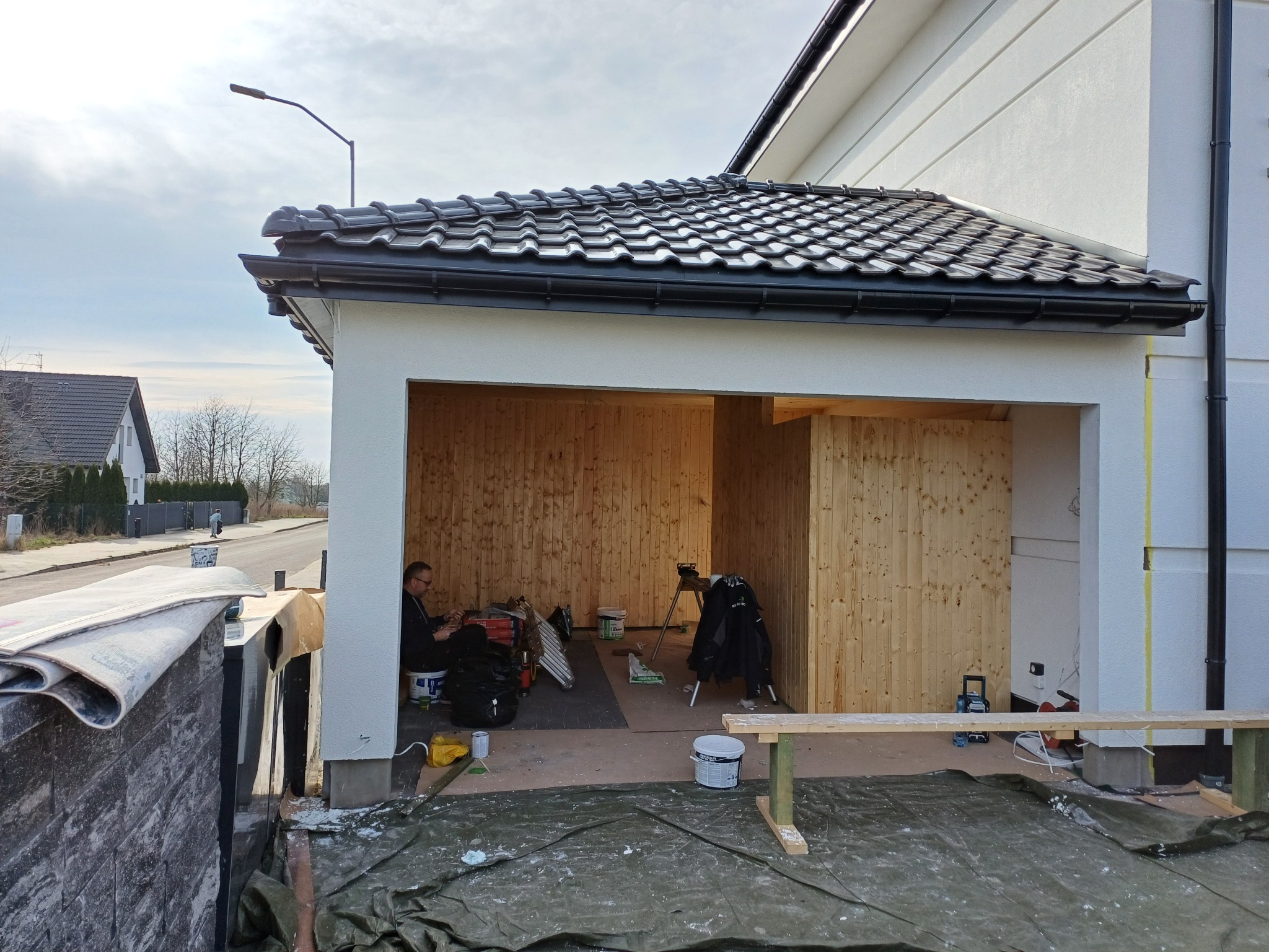 Prace wykończeniowe przy nowym dachu z czarnej dachówki i orynnowaniu. W tle widoczny pracownik oraz narzędzia na placu budowy.