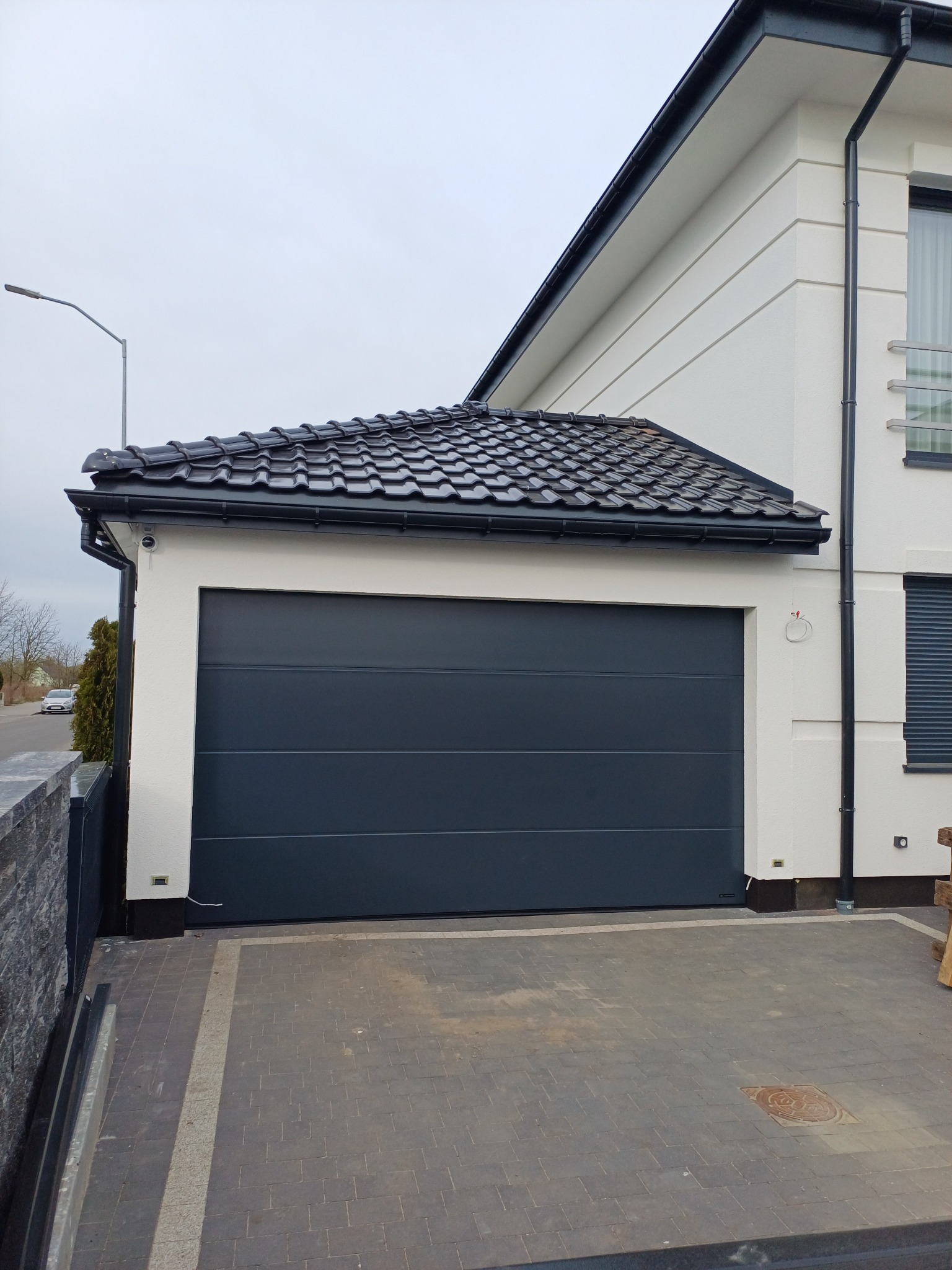 Nowoczesny garaż z ciemną bramą segmentową i czarnym dachem z dachówki ceramicznej, widok z zewnątrz na tle jasnej elewacji budynku.