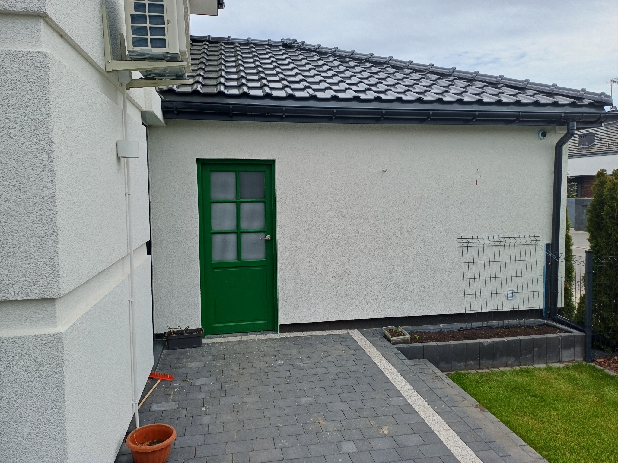 Biały budynek z ciemną dachówką i zielonymi drzwiami, widok z boku, kostka brukowa na pierwszym planie, rynna wzdłuż dachu, fragment ogrodu z tyłu.