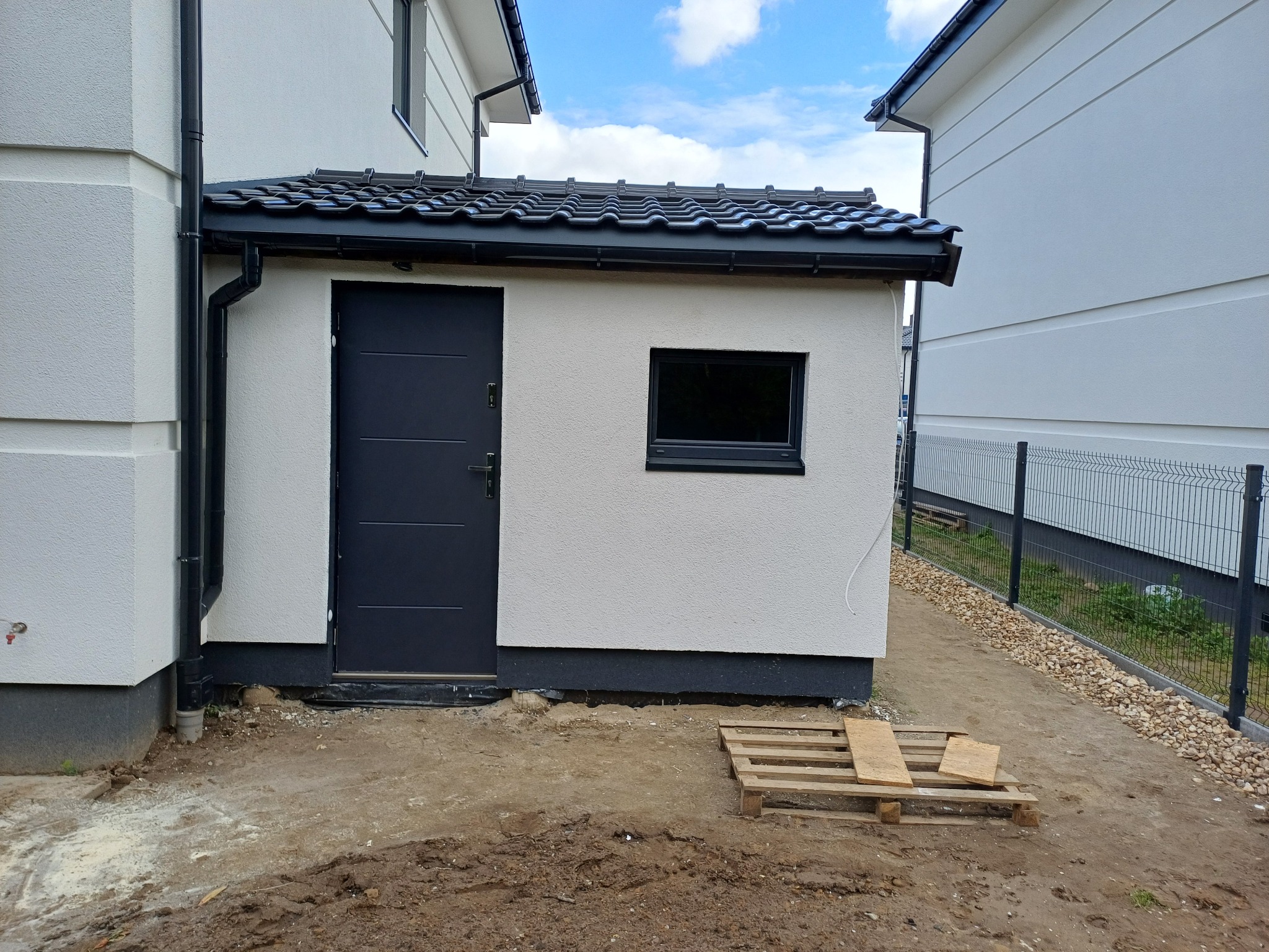 Nowoczesny, biały budynek gospodarczy z czarnym dachem i drzwiami, widoczny z boku na tle błękitnego nieba. Paleta drewniana leży na ziemi.