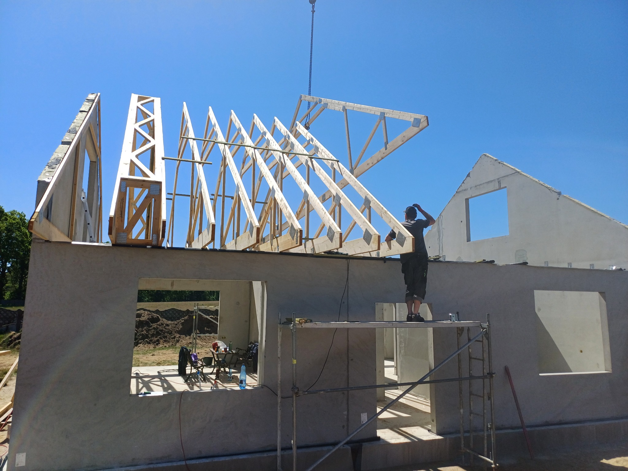 Montaż drewnianej więźby dachowej na budynku z betonu komórkowego, widok z rusztowania na pracownika mocującego element konstrukcji za pomocą dźwigu.