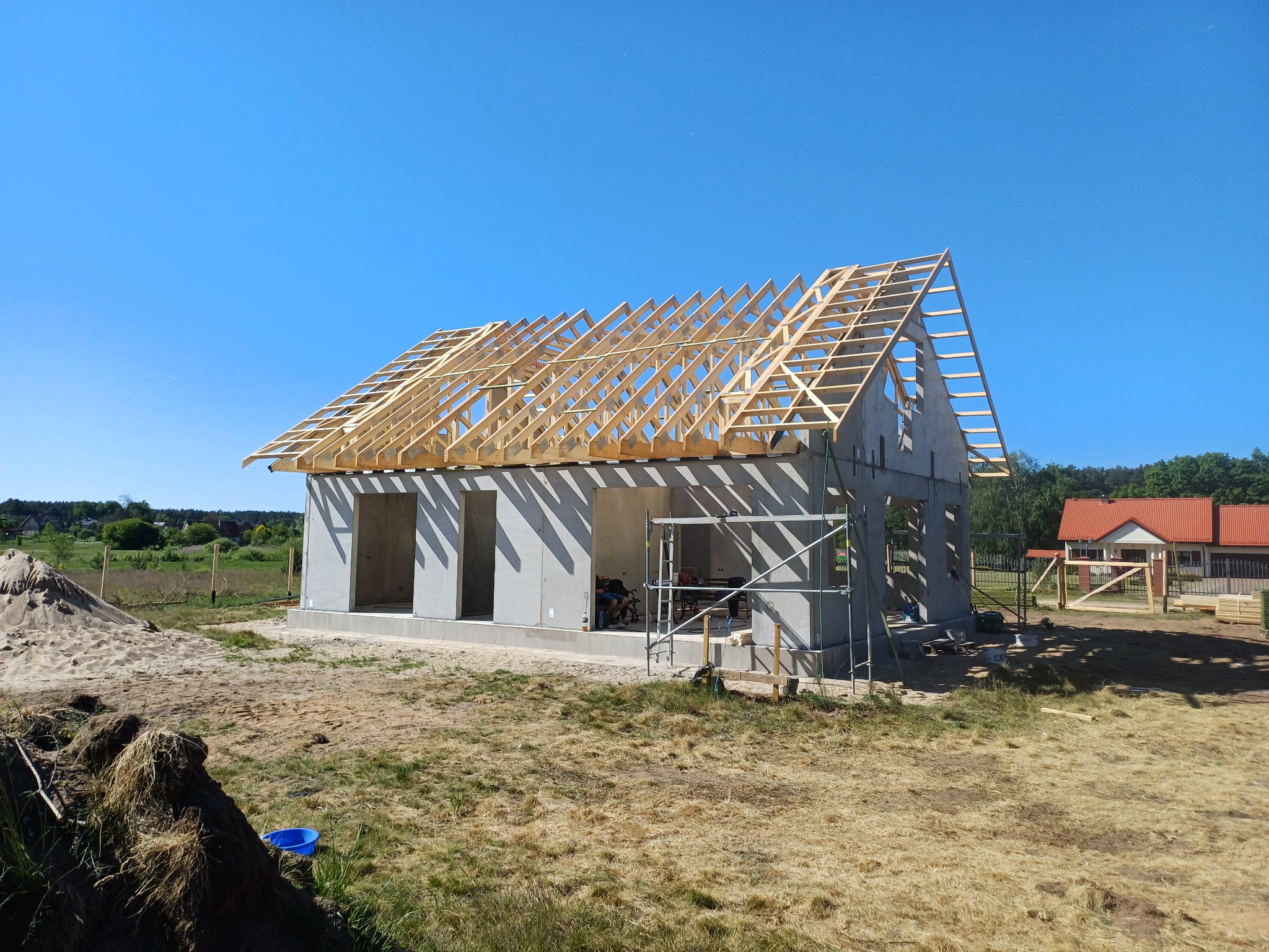 Surowy dom z betonu komórkowego w trakcie budowy, z drewnianą konstrukcją dachu w słoneczny dzień. Widoczne rusztowania i teren budowy.
