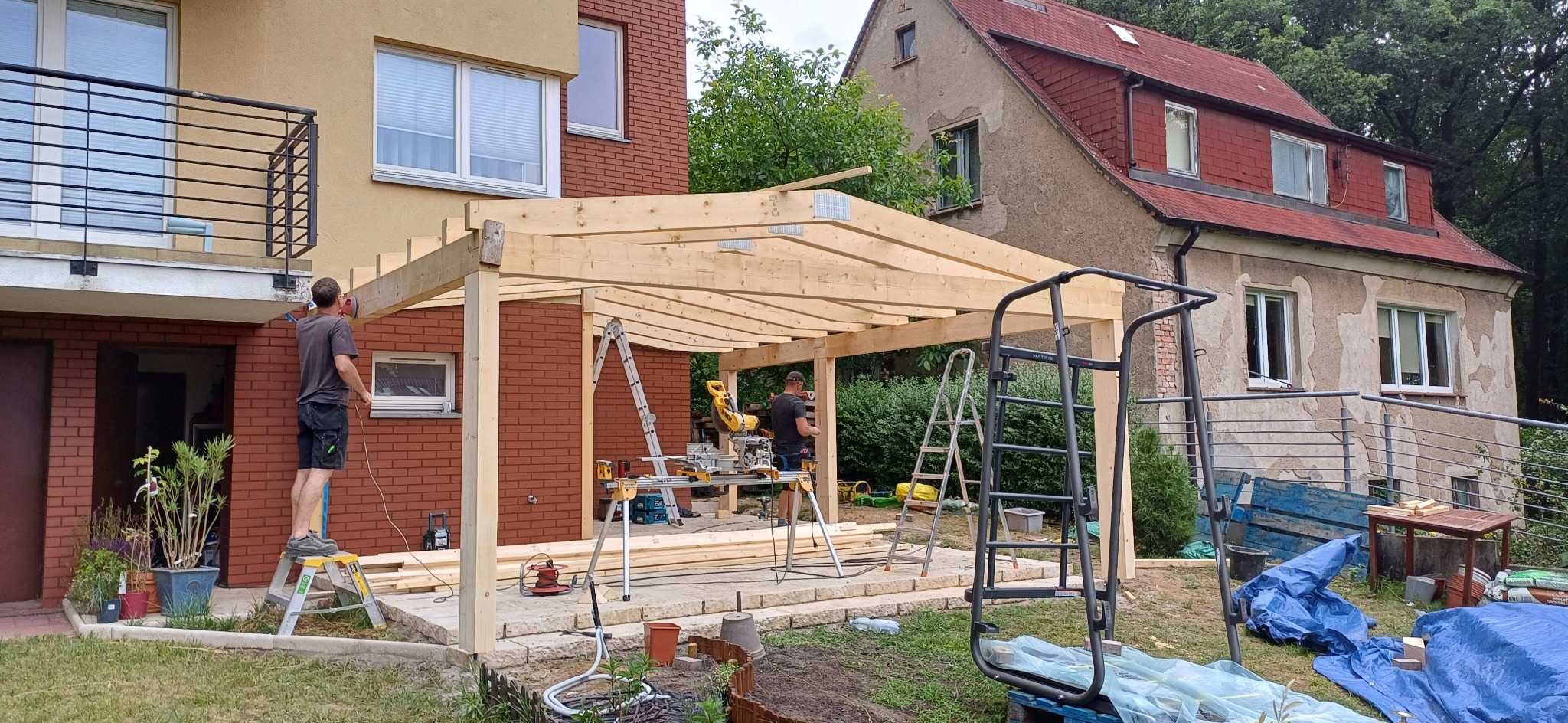 Budowa drewnianej pergoli przy domu z cegły. Dwóch mężczyzn pracuje przy konstrukcji, używając narzędzi. W tle widoczny dom z czerwonym dachem.