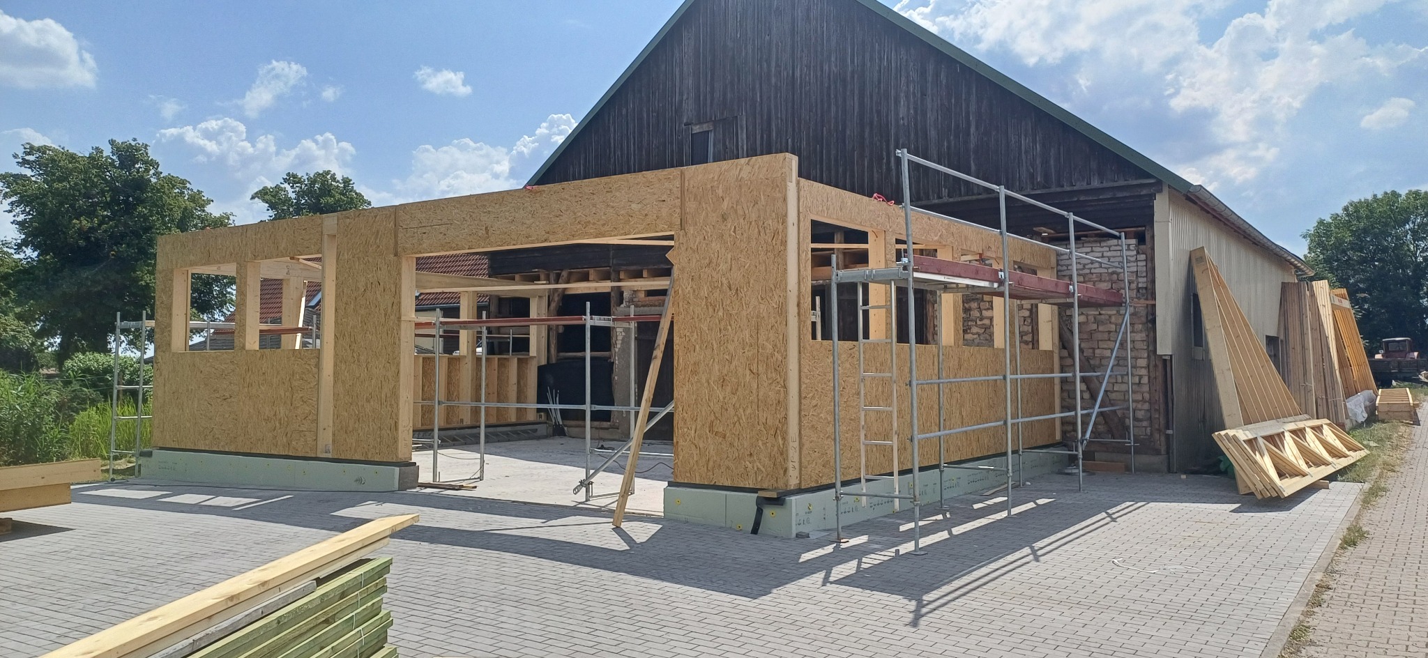 Budowa szkieletowa budynku w trakcie realizacji, z widocznym drewnianym szkieletem i płytami OSB, rusztowaniem oraz elementami konstrukcyjnymi na zewnątrz.