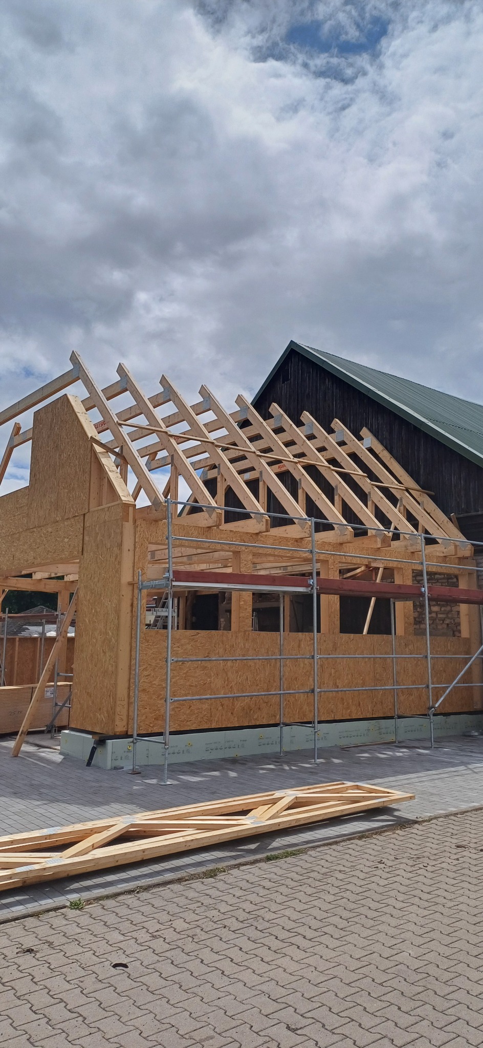 Wznoszenie drewnianej konstrukcji domu z widoczną więźbą dachową i rusztowaniami na tle pochmurnego nieba. Częściowo widoczny budynek w tle.