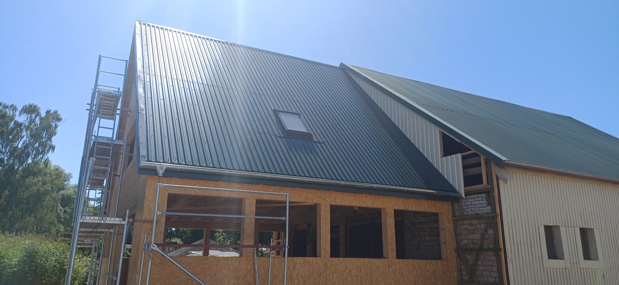 Montaż dachu z blachy trapezowej z oknem dachowym, widok na budynek w trakcie budowy z rusztowaniem i niezabudowanymi otworami okiennymi.