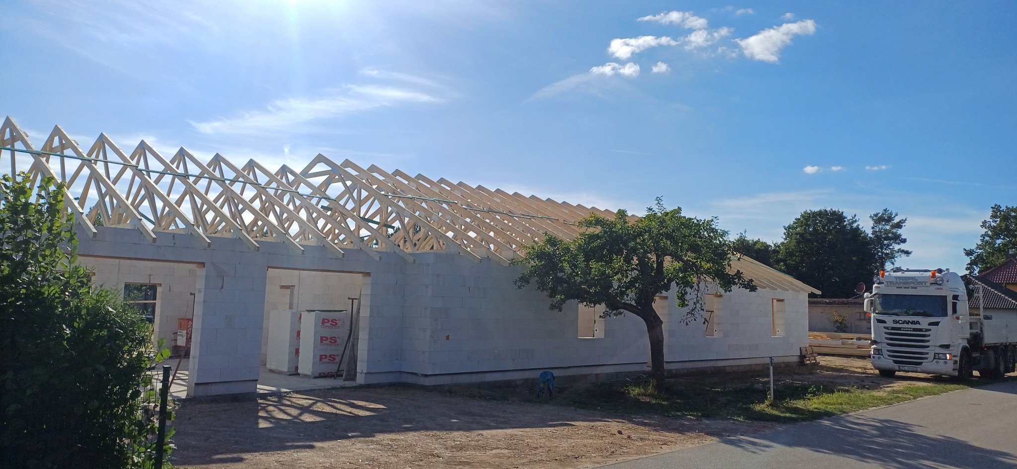 Wznoszenie więźby dachowej na budynku z bloczków betonu komórkowego, widok z zewnątrz, drzewo w pobliżu, ciężarówka w tle, słoneczny dzień.