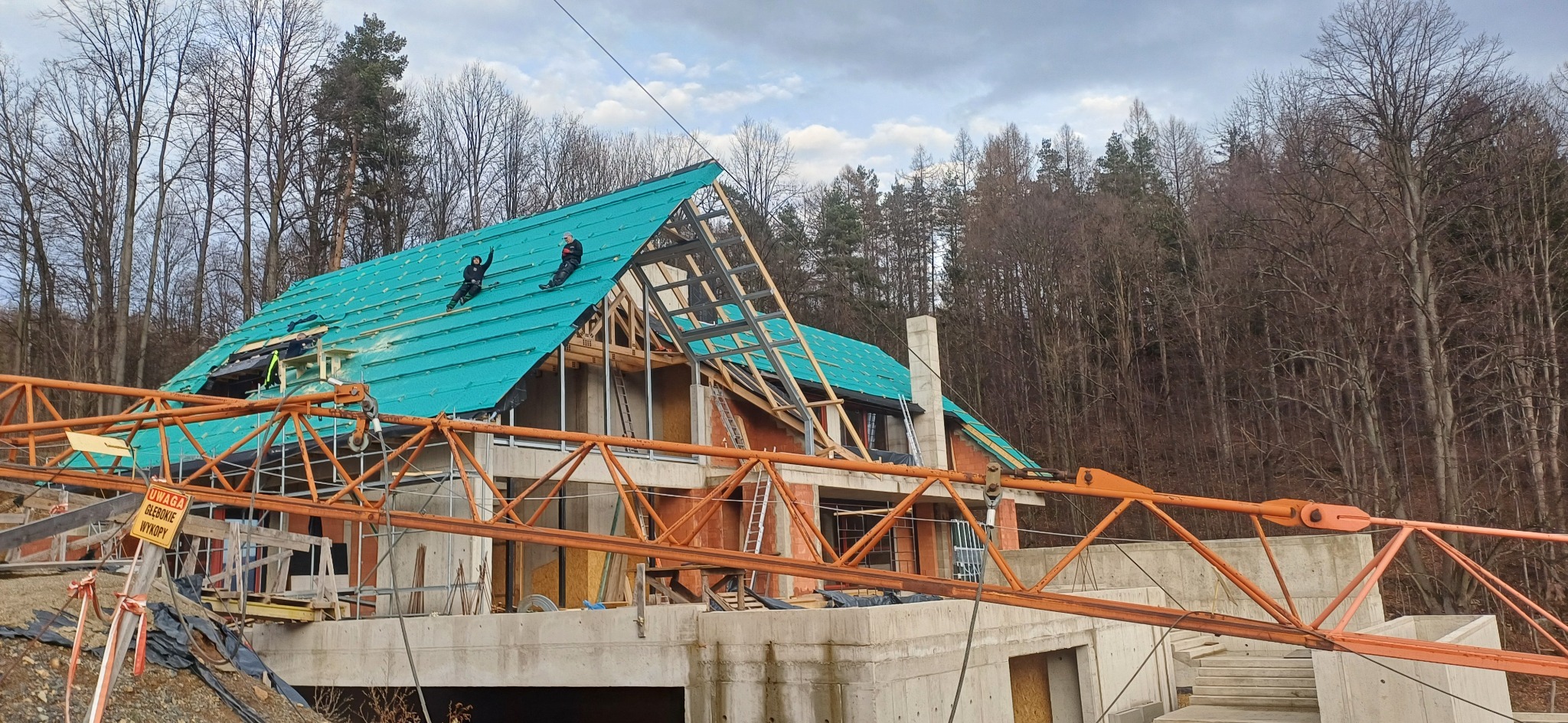 Budowa domu jednorodzinnego z zielonym dachem w trakcie realizacji. Na dachu dwóch pracowników. Na pierwszym planie pomarańczowy dźwig budowlany.