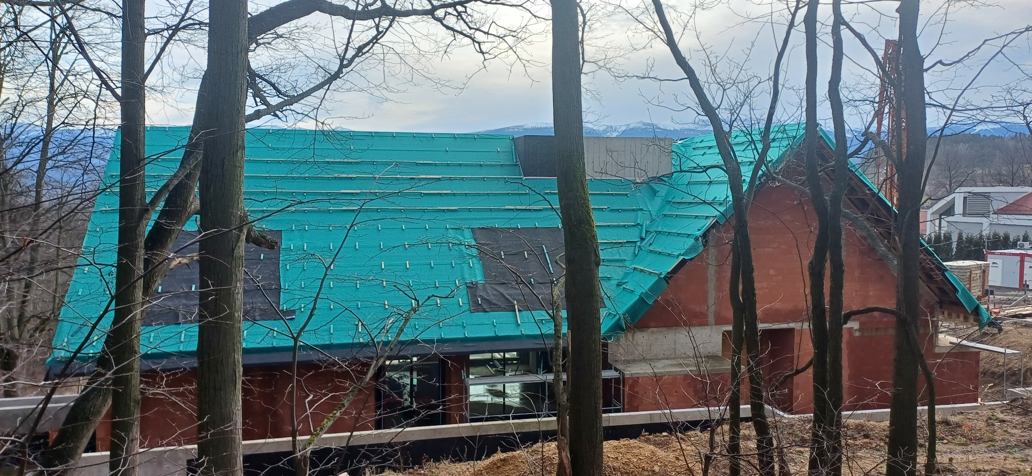 Dom w trakcie budowy z widocznym dachem pokrytym membraną dachową w kolorze turkusowym, częściowo zabezpieczonym czarną folią, na tle gór i drzew.