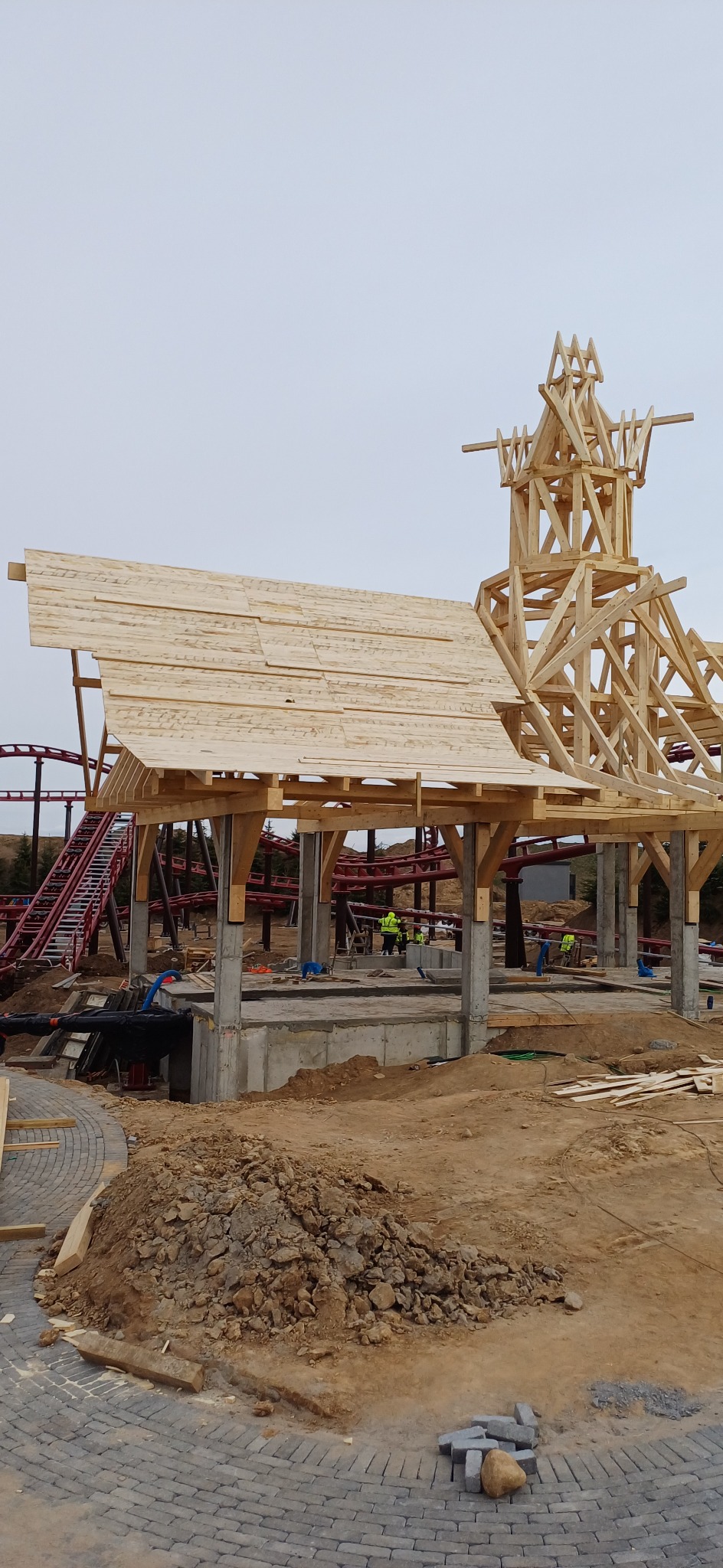 Drewniana konstrukcja dachu w trakcie budowy, z elementami więźby dachowej i fragmentem kolejki górskiej w tle, na placu budowy z ziemią i kostką brukową.