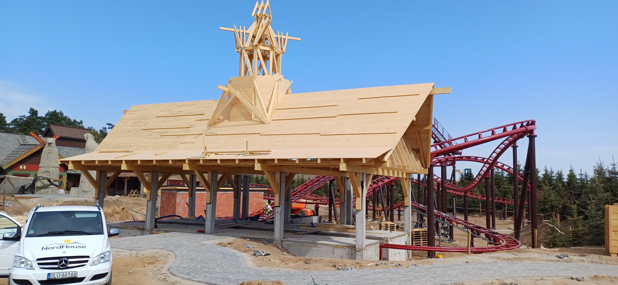 Drewniana konstrukcja dachu z wieżyczką w stylu skandynawskim, w tle czerwony rollercoaster i budynki. Widoczny samochód dostawczy z logo firmy.